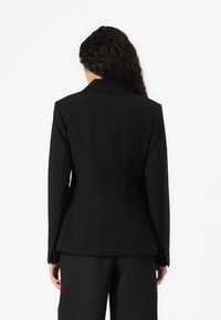 Blazer noir ajusté avec une forme structurée, épaules marquées et texture lisse ; présente une fente au dos et des poignets boutonnés.