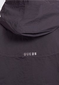 Czarna kurtka z kapturem wykonana z lekkiego materiału, z błyszczącym srebrnym logo „GUESS” na plecach oraz subtelnymi detalami tekstury.