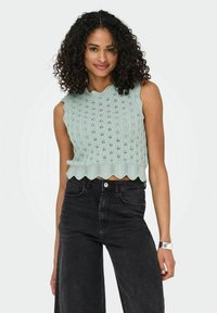 Lichtgroen gebreid crop top met gekartelde randen en een open steekontwerp, gecombineerd met hoge zwarte wijde jeans. Zilveren horloge om de pols.