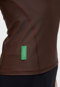 Gros plan d'une chemise marron ajustée avec des coutures visibles et une petite étiquette en tissu verte indiquant "District Vision" près de la couture latérale.