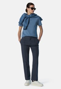 T-shirt in cotone blu con collo a giro, abbinata a pantaloni navy con cuciture laterali. Sneakers bianche e un maglione blu appoggiato sulle spalle.