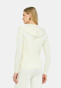 Maglione crema con cappuccio e texture a treccia sulla schiena, design attillato, polsini a coste e un piccolo accento dorato vicino al collo.