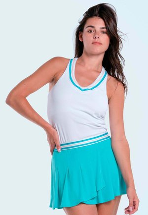 Joven mujer con top blanco sin mangas con ribete azul y falda plisada turquesa, posando con la mano en la cadera contra un fondo claro.