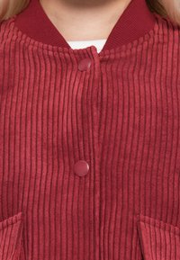 Close-up of red ribbed jacket with two round buttons and a collar over a white top.