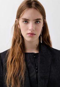 Blazer noir texturé avec un col structuré, présentant des boutons décoratifs et de longs cheveux bruns ondulés. Des boucles d'oreilles pendantes en argent rehaussent le look.