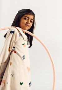 Abito color crema con un colletto arruffato, caratterizzato da vari motivi colorati come frutti e animali. Un hula hoop rosa è appoggiato attorno a chi lo indossa.