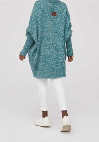 Gruby sweter w kolorze teal z fakturowanym wykończeniem, w oversize'owym kroju, z długimi rękawami i brązową łatą z logo na plecach. W komplecie z białymi spodniami.