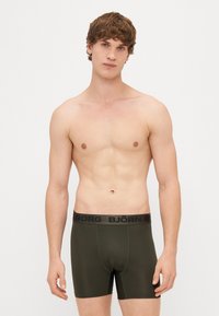 Fiatal férfi göndör hajjal, sötétzöld Björn Borg boxer alsóneműt visel, egy sima fehér háttér előtt áll.