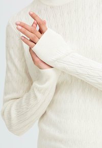 White cable-knit sweater with ribbed cuffs. The texture features a repeating pattern of twisted cables, offering a layered, tactile appearance.