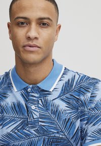 Blå polo skjorta med palmbladstryck i mörkblått, med en vit krage och knappar. Inkluderar broderad logodetalj på bröstet.
