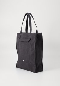Black tote bag made from textured fabric, featuring two sturdy handles, a front pocket, and a minimalistic logo at the bottom.