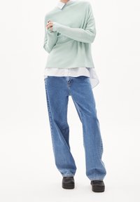 Pull vert menthe clair superposé sur une chemise blanche à col, associé à un jean bleu clair ample et des sabots noirs. Textures lisses, style décontracté.