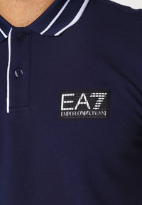 Marinblå polo-shirt med vitrandig krage, tvåknappsslutning och en rektangulär EA7 Emporio Armani-logotyp på bröstet.
