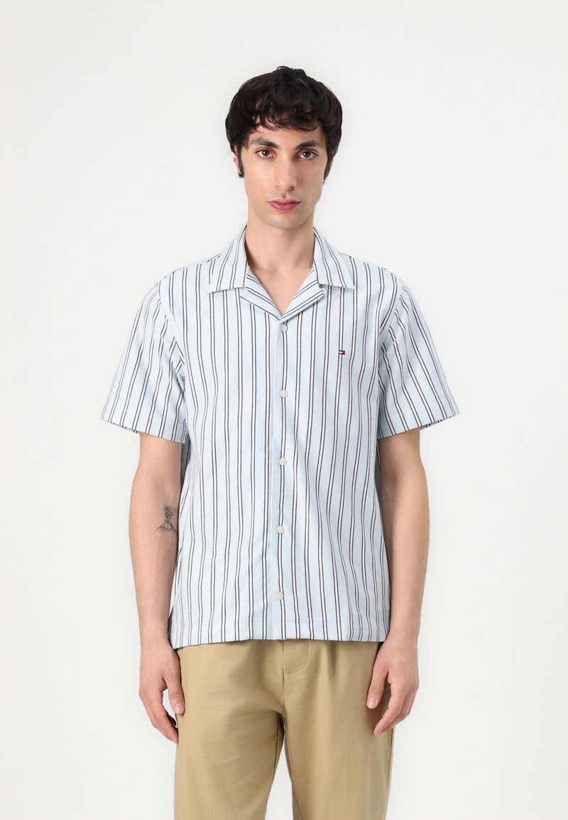 Camisa de botones de manga corta en azul claro con rayas verticales oscuras, hecha de tela suave. Tiene un cuello tipo camp y cierre de botones en la parte frontal.