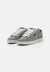 Sive sneakers od antilopa s bijelim vezicama, niskim profilom, crnim akcentima i logom na jeziku i bočnoj strani.