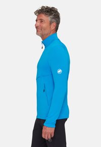 Niebieska polarowa kurtka zapinana na zamek z wysokim kołnierzem, długimi rękawami i kieszenią zapinaną na zamek z boku. Posiada logo na lewym rękawie. Gładka tekstura.