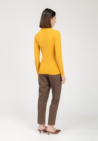 Gelber, figurbetonter Pullover mit hohem Kragen, kombiniert mit braunen, schmalen Hose und braunen, Absatzmules, von hinten gezeigt.