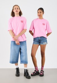 Zwei Personen tragen passende, übergroße pinkfarbene T-Shirts mit einem Logo. Die eine kombiniert es mit Denim-Culottes, die andere mit zerrissenen Denim-Shorts und schwarzen Sandalen.