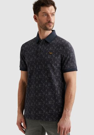 Mężczyzna w ciemnej, wzorzystej koszulce polo z kołnierzykiem i trzema guzikami, stojący i patrzący na bok na tle jednolitego tła.