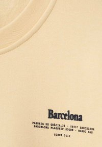 Beige textiel met een ronde halslijn voorzien van zwarte gedrukt tekst: "Barcelona," adresgegevens en "Mango Man" branding onderaan.