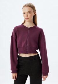Sudadera con cremallera recortada en tejido púrpura profundo, con mangas largas, dobladillo acanalado y un diseño minimalista. Combinada con pantalones negros.