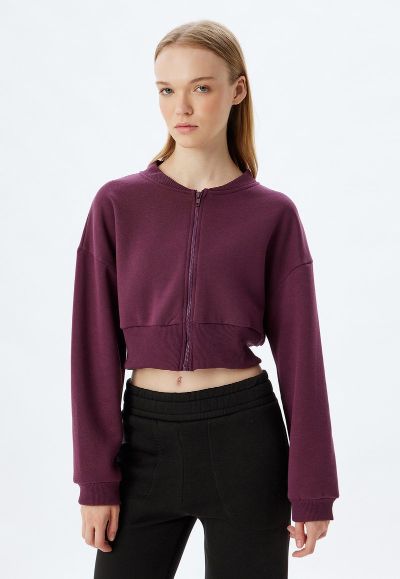 Sudadera con cremallera recortada en tejido púrpura profundo, con mangas largas, dobladillo acanalado y un diseño minimalista. Combinada con pantalones negros.