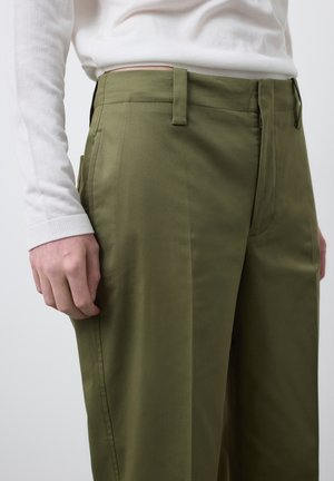 Gros plan sur une personne portant un pantalon vert olive et un haut à manches longues blanc, montrant la poche latérale et les passants de ceinture du pantalon.