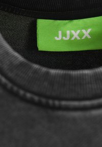 Tejido negro con un cuello redondo; presenta una etiqueta verde con "JJXX" en texto blanco. Costura interna texturizada visible; superficie suave.