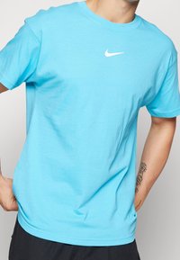 Hombre con una camiseta de Nike azul claro de mangas cortas, pantalones negros y un tatuaje visible en el antebrazo izquierdo.