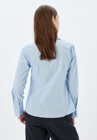 Camisa de botones azul claro con mangas largas, ajuste entallado y detalles en los puños, combinada con pantalones a rayas. Textura de tela suave y lisa.