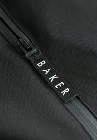 Schwarze Stofftasche mit Reißverschluss. Ein kontrastierendes weißes Etikett zeigt das Wort "BAKER". Weiche Textur mit einem klaren, minimalistischen Design.