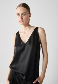 Top senza maniche in satin nero con scollatura a V, caratterizzato da una texture liscia e una vestibilità rilassata. Accentuatato da spalline sottili e pieghe laterali.