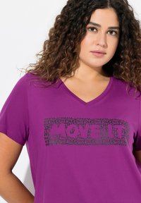 Lilla kortærmet V-hals t-shirt med en fed sort grafik med gentagne textelementer "MOVE" og "POWER." Glat stofstruktur.