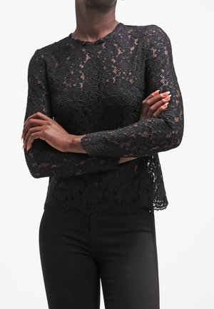 Kobieta ubrana w czarną, długorękawową koronkową bluzkę w kwiaty z falowanym brzegiem, ze skrzyżowanymi rękami, w połączeniu z czarnymi dopasowanymi spodniami.