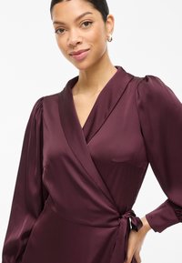 Φόρεμα σε στυλ wrap από σατέν μπορντό, με μακριά φουσκωτά μανίκια, βαθύ V-λαιμόκοψη και λεπτομέρεια δεσίματος στο πλάι. Λείο υλικό και διακριτική λάμψη τονίζουν το σχέδιο.