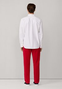 Persona de pie de espaldas, vistiendo una camisa blanca de manga larga, pantalones rojos y zapatos negros sin cordones contra un fondo liso.