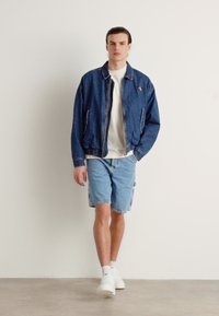 Giacca di jeans con chiusura a zip, T-shirt blu chiaro e pantaloni corti coordinati blu, abbinati a sneakers bianche. Tessuto testurizzato, vestibilità casual.