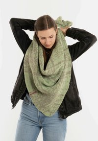 Zielony, wzorzysty sweter w formie szalika owinięty wokół szyi, z gradientowym wzorem. Towarzyszy mu czarna kurtka, z widoczną metką.