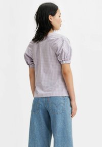 Camicia a maniche corte a quadri viola con maniche a sbuffo, abbinata a jeans a gamba larga blu. La camicia presenta un bottone alla manica.