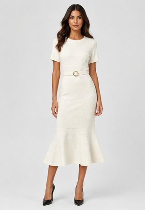 Woman wearing a fitted off-white midi dress with short sleeves, textured dots, a belt with a gold buckle, and flared hem, paired with black heels.