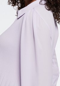 Ljuslila blus med en texturerad, plisserad tyg. Har en klassisk krage och voluminösa ärmar som är samlade vid axeln.