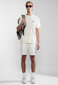Crèmekleurig T-shirt met blauwe grafische print, witte shorts, grijze rugzak, witte sneakers, zonnebril en roestvrijstalen waterfles.