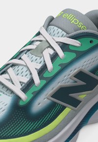 Αθλητικό παπούτσι με λευκά κορδόνια, μπλε και πράσινο πλέγμα επάνω μέρος, νέον κίτρινα στοιχεία και μεγάλο λογότυπο "N" στο πλάι.