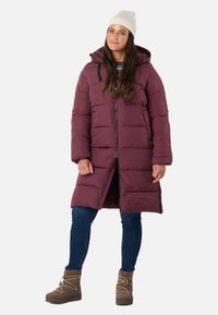 Kvinde iført en lang mørkerød boblejakke, blå jeans, beige vinterstøvler og en hvid strikket hue, stående med hænderne afslappet langs siden.
