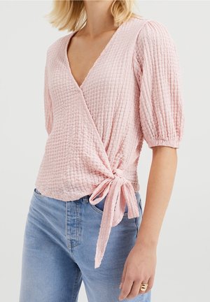 Haut cache-cœur rose en tissu texturé froissé, col en V, manches courtes bouffantes et détail de nouage sur le côté. Associé à un jean bleu clair.