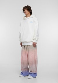 Felpa bianca con tasca frontale e logo blu; abbinata a pantaloni larghi con una sfumatura pastello, che passa dal rosa al blu ai polsini.