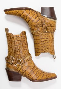 Botas de tobillo de cuero amarillo-marrón con textura de cocodrilo, punta afilada, cuello con picos y detalles de hebilla en el costado. Tacón de madera.