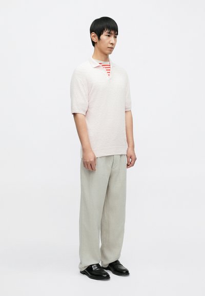 Ροζ ανοιχτού χρώματος πόλο μπλούζα με υφή, κοντά μανίκια, φορεμένη πάνω από κόκκινο και λευκό ριγέ πουκάμισο. Συνδυασμένη με άνετο ανοιχτό γκρι παντελόνι και μαύρα παπούτσια.