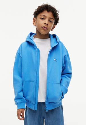 Dreng med krøllet hår iført en blå hættetrøje med lynlås over en hvid t-shirt og blå jeans, står med hænderne i lommerne.