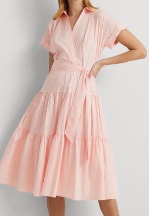 Kobieta ubrana w jasnoróżową, krótko rękawową, warstwową sukienkę kopertową z kołnierzem i wiązanym paskiem w talii, stojąca na jednolitym tle.